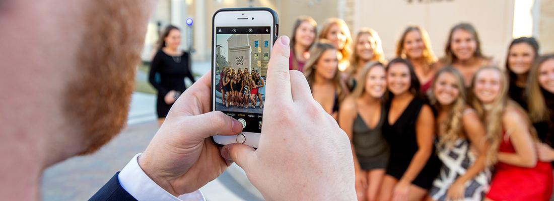 在TCU希腊村的派对前，一个年轻人用手机拍摄一群微笑的姐妹会女孩.